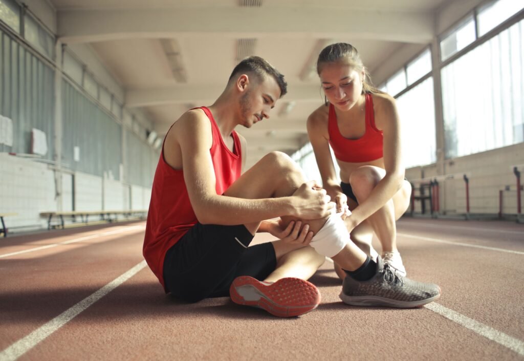 Side view of athlete sitting on rubber track in sports hall while sportive female partner helping to band leg after injury