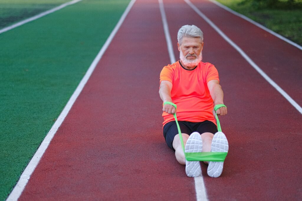 Full body of diligent elderly male with beard warming up with elastic rubber on racetrack in stadium