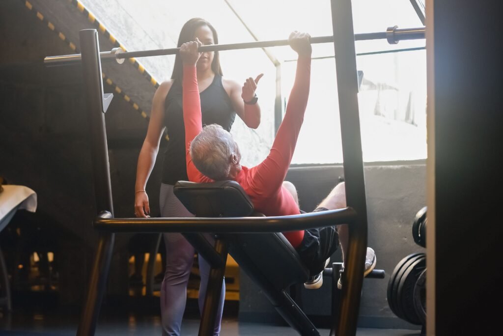 Senior man lifting weights in a gym with guidance from a female trainer, promoting fitness and a healthy lifestyle.